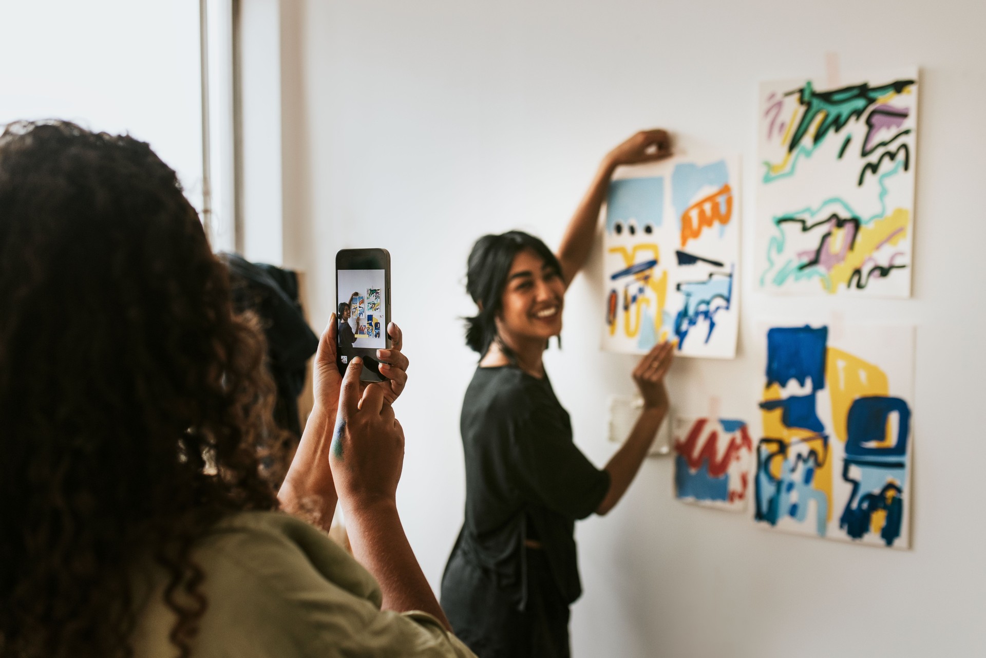 Friend taking a photo with phone camera of her artist friend by a mood board on a white wall